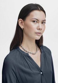 Zilveren ketting, grijze blouse met textuur en een geplooide kraag met knoopdetails. Model heeft lang donker haar en kleine creolen.