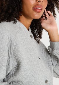 Sudadera gris texturizada con un patrón acolchado, puños acanalados y dos botones de presión negros en la parte frontal. Tela suave y acogedora.