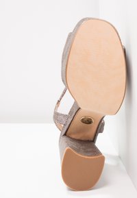 Escarpin beige à talon haut avec un dessus en maille texturée, une bride de cheville adjustable et une semelle lisse de couleur claire présentant un motif de semelle.