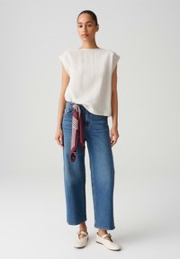 Witte linnen top met korte mouwen, hoge halslijn, gecombineerd met blauwe wijdlopende jeans en een bordeauxrode gestreepte zijden sjaal die om de taille is geknoopt.