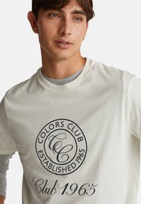 Světlý béžový tričko s kulatým logem v tmavě modré barvě se nápisem "Colors Club" a "Established 1965." Kulatý výstřih a měkká textura.