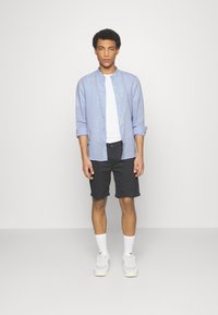Camisa de lino azul claro con mangas largas, camiseta blanca de tirantes, pantalones cortos negros, calcetas blancas y deportivas grises. Modelo de pie frente a un fondo liso.