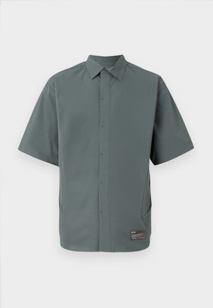 Kortærmet skjorte i dæmpet grøn stof, med en klassisk krave, knaplukning foran, sidelommer og et mærke ved sømmen.