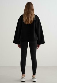 Fekete, túlságosan nagy bolyhos pulóver széles ujjal, fekete leggings-szel viselve, fehér sportcipőkkel párosítva. Háttal áll egy semleges háttér előtt.