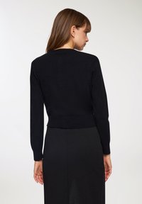 Maglione nero lavorato a maglia con maniche lunghe, orlo a coste e scollatura rotonda, indossato sopra una gonna nera. Texture morbida, design aderente.