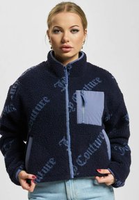 Ženska oblečena v mornarsko modro flis jakno z svetlo modrim zadrgo, žepom in ponovljenim vzorcem besedila, v kombinaciji s svetlo modrimi jeans hlačami.