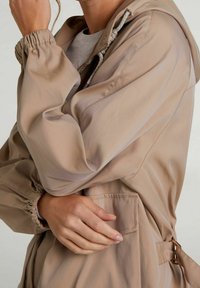 Beżowa lekką kurtkę z kapturem, elastycznymi mankietami i paskiem w talii. Posiada gładką teksturę i dekoracyjne sznureczki przy kołnierzu.