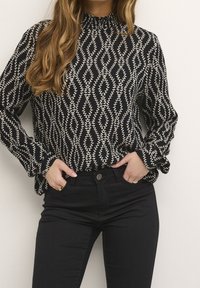 Blouse à motifs noir et blanc avec un col montant, des manches longues et un motif de feuilles tourbillonnantes. Associée à un jean noir ajusté.