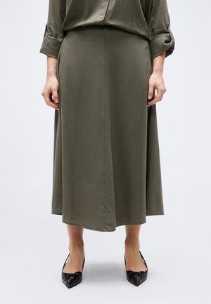 Kvinde iført en olivengrøn midinederdel og matchende skjorte med knapper og opkørt ærmer, kombineret med sorte spidse høje hæle.