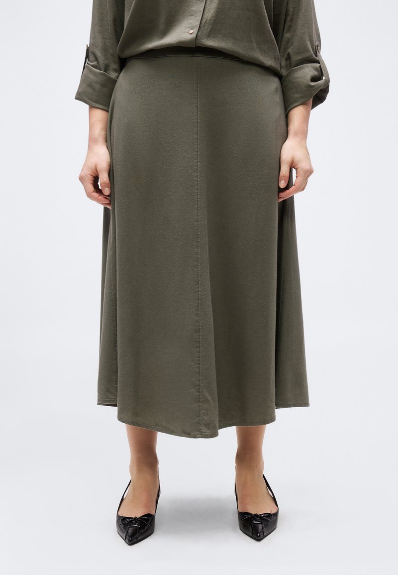 Mujer con falda midi verde oliva y camisa a juego abotonada con mangas remangadas, combinadas con tacones negros de punta afilada.