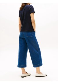 Granatowa koszulka z krótkim rękawem w połączeniu z szerokimi ciemnoniebieskimi dżinsami. Dżinsy mają mankiety na końcach nogawek oraz dwie tylne kieszenie. Całość dopełniają białe mokasyny.