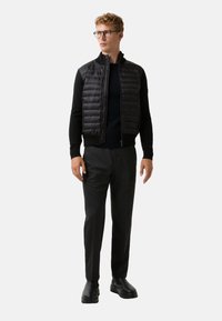 Czarna kamizelka puchowa z zapięciem na zamek, wysokim kołnierzem i teksturowanym materiałem, noszona na czarnym swetrze, w zestawieniu z ciemnymi spodniami i czarnymi butami.