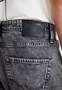Black-gray denim jeans with a textured surface, featuring a leather logo patch on the waistband and subtle stitching on the back pockets.
