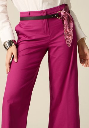 Pantalones anchos magenta con botón y cremallera, cinturón negro con anillo dorado y pañuelo rosa con estampado atado a la cintura, combinados con blusa blanca.