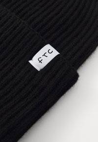 Černá pletená čepice s ribbovanou strukturou a bílou nášivkou, na které je černými písmeny vyšitý nápis "ftc" na boku.
