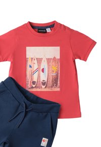 Camiseta de algodón roja con un gráfico de tabla de surf, combinada con pantalones de chándal azul marino que presentan un logo bordado. Mangas cortas, corte casual.