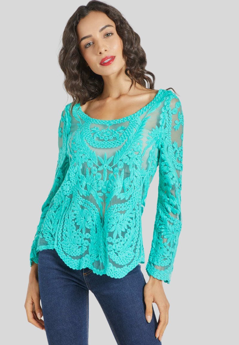 Mujer con cabello oscuro y ondulado, vistiendo una blusa de encaje turquesa transparente de manga larga con patrones florales y jeans azul oscuro, de pie contra un fondo gris.