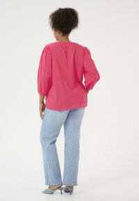 Blouse rose à manches bouffantes avec des points texturés, encolure arrière avec une petite ouverture, associée à un jean droit bleu clair et des sandales à talons bleues.