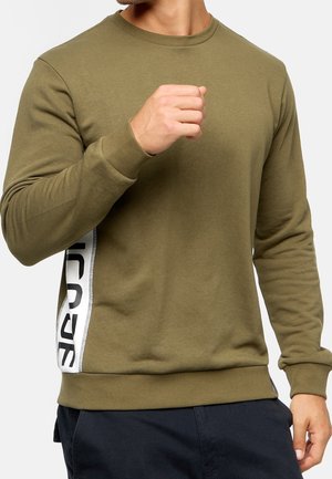 Hombre vistiendo sudadera de manga larga verde oliva con texto vertical en blanco y negro en el costado, levantando el puño, recortado a la altura del cuello y la cintura.