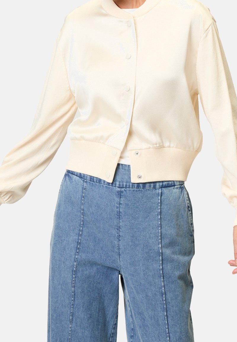 Femme portant un cardigan beige clair boutonné avec une taille côtelée et un jean taille haute à jambes larges bleu clair sur fond uni.