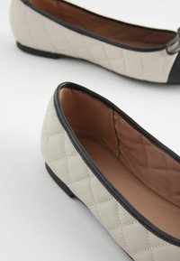 Gequiltete beige Ballerinas mit schwarzen Akzenten, flache Sohle und glattem Innenfutter, mit dekorativer Naht und abgerundetem Zehenbereich.