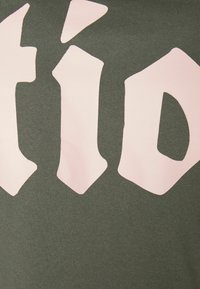 Zelený bavlněný oděv s velkými, nepravidelnými růžovými písmeny. Textura vypadá měkce s hladkým povrchem a výrazným barevným kontrastem.