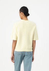 Lys gul kortermet genser med myk, teksturert strikk. Har en bred rund hals og rett kant, brukt med lyseblå jeans.