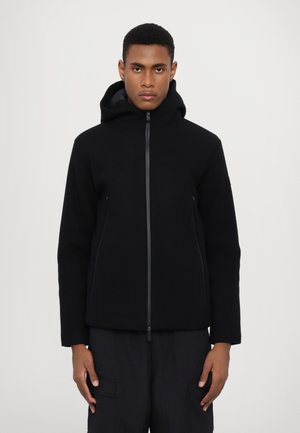 Giacca nera con cappuccio e zip frontale, realizzata in materiale morbido. Presenta due tasche laterali e una silhouette aderente. Abbinata a pantaloni scuri.