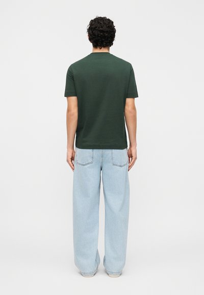 Άνδρας με πλάτη φοράει σκούρο πράσινο κοντομάνικο πουκάμισο και ανοιχτό γαλάζιο φαρδιά παντελόνι τζιν μπροστά σε απλό λευκό φόντο.