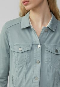  Lichtblauwe denimjas met een kraag, twee borstzakken en zilveren drukknoopjes. Gladde textuur met zichtbare stiksels.