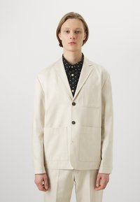 Beige bomullskavaj med två framlommor, enkelknappslåsning och snibbkrage, bärs över en svart prickig skjorta. Slät textur.