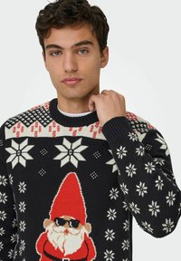 Joven con un suéter navideño negro con copos de nieve y un Santa cool con gafas de sol, sosteniendo el cuello del suéter.