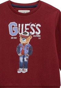 Felpa bordeaux con grafica di un orso che indossa un berretto da baseball, una giacca di jeans e dei jeans. Include il logo "GUESS" e l'anno di fondazione.