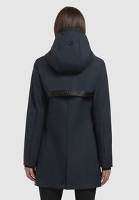 Marinblå vattentät jacka med huva, utrustad med en logotypförsedd rem över ryggen, långa ärmar och rak skärning.