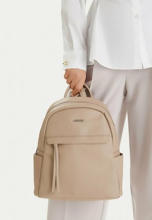 Mano che tiene uno zaino in pelle beige Lasocki dai manici superiori, indossato con camicia bianca e pantaloni chiari.