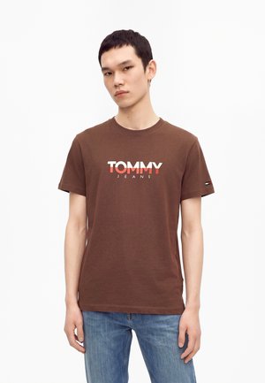 Млад мъж, носещ кафява тениска Tommy Jeans и сини дънки, стои пред бял фон.
