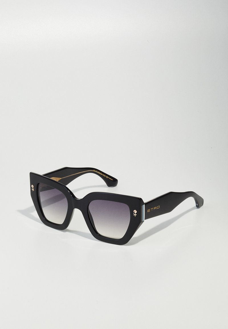 Black eyeglasses with a bold, angular cat-eye shape. Gradient lenses transition from gray to clear. Gold accents and logo on the temples.