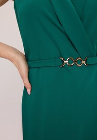Sukienka w kolorze szmaragdowym z złożonym wzorem, wąskim pasem ozdobionym złotym okrągłym akcentem oraz gładką fakturą materiału.