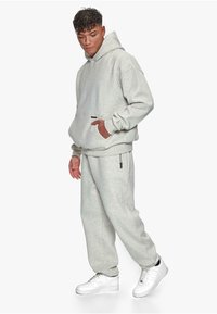 Light grey hoodie with a front pocket and adjustable hood, paired with matching trousers featuring a side zip pocket. White sneakers.