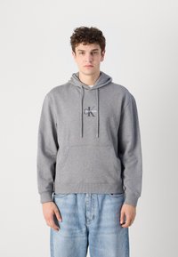 Pilkas džemperis su kapuce, pagamintas iš medvilnės ir poliesterio mišinio. Jame yra priekinė kišenė, virvutė ant kapučio ir mažas Calvin Klein logotipas ant krūtinės.