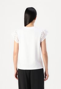 T-shirt en coton blanc avec des manches bouffantes courtes en œillets, col rond et coupe droite au niveau de l'ourlet, porté avec un pantalon noir.