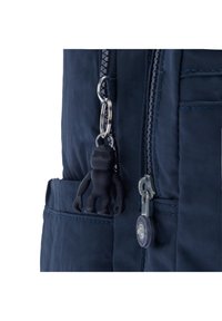 Sac à dos en nylon bleu marine avec une poche zippée, un porte-clés en caoutchouc en forme de singe, et des accessoires en métal. Tissu texturé avec des détails de couture.