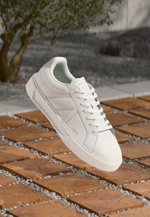 Baskets basses blanches avec le logo « ARKK », placées au-dessus d'un sol en bois et galets à motifs, avec un arbre et un fond en pierre.