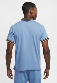 Lys blå kortermet polo-skjorte med teksturert stoff, brun og hvit stripete krage og rett hem, sett fra baksiden.