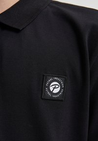 Svart polo t-shirt i texturerad tyg, med en fyrkantig svart patch med vit "Petrol Industries" logotyp på vänster bröstområde.
