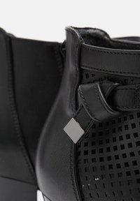 Black ankle boots with perforated leather, featuring a flat heel and a textured square accent on the strap. Smooth finish, modern design.