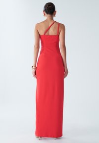 Punainen, lattiaan ulottuva mekko, jossa on vartalonmyötäinen yläosa, olkapään yli kulkeva hihna ja virtaviivainen siluetti. Tasainen, kevyt kangas, vähäiset koristeet.