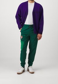 Lila kofta med knappar, vit t-shirt, gröna träningsbyxor med sidofickor och ett Portugal-emblem, kombinerat med vita och bruna sneakers.