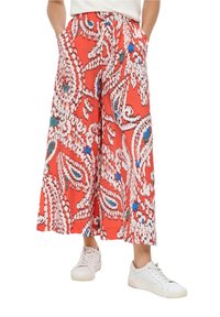 Rote weite Hose mit einem weißen und blauen Paisleymuster, ausgestattet mit einem elastischen Bund und seitlichen Taschen. Wird mit weißen Turnschuhen getragen.