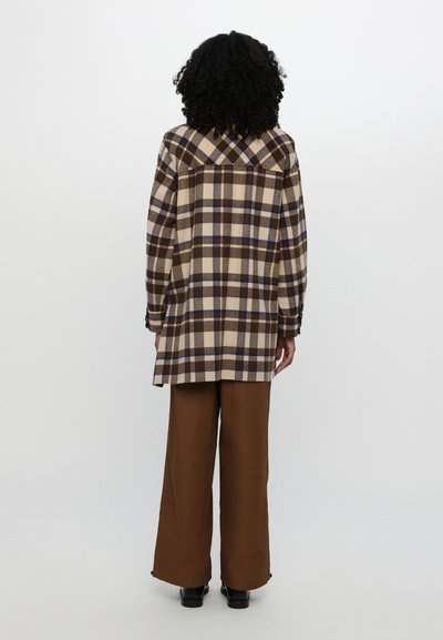 Camisa de cuadros marrón y beige con acentos azules, de mangas largas y corte holgado, combinada con pantalones anchos marrones y zapatos negros.